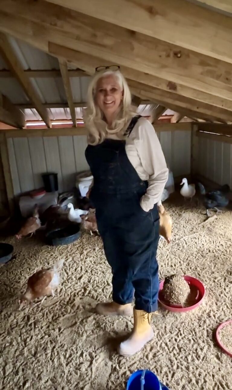 Jules Morris in denim overalls and work boots standing on fresh pine shavings inside the large coop, hens and white ducks feeding around her