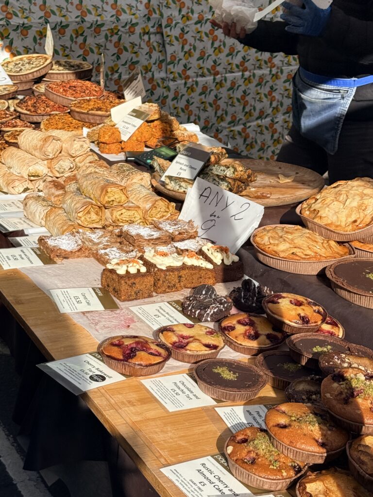 Market stall counter covered in fruit tarts, quiches, chocolate tarts and sausage rolls with handwritten price cards