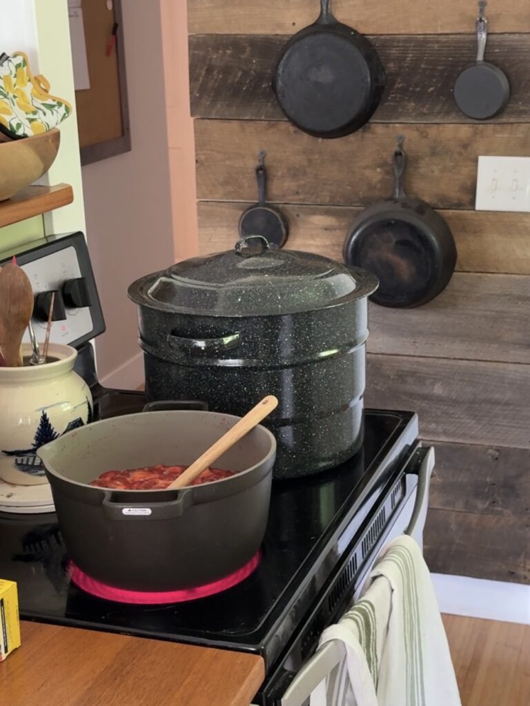 Pot simmering on the stovetop with a wooden spoon and a large canner behind it, cast iron hanging on the reclaimed wood wall