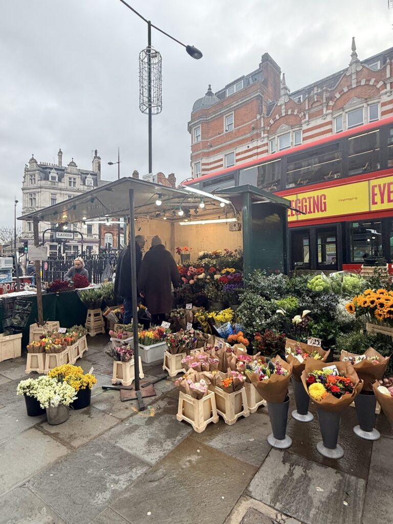 Outdoor London flower stall filled with wrapped bouquets and buckets of blooms, a red double decker bus passing behind