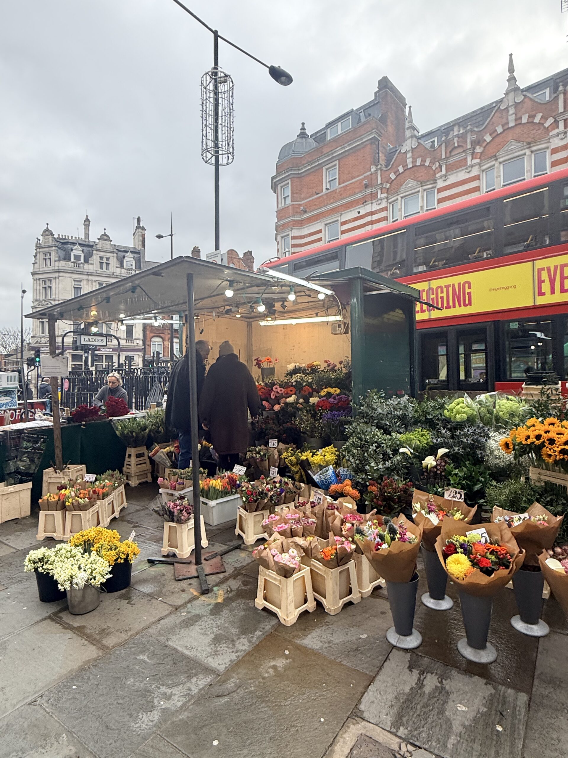 Outdoor London flower stall filled with wrapped bouquets and buckets of blooms, a red double decker bus passing behind