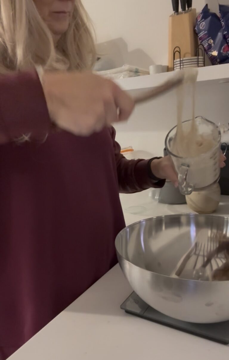 Jules Morris pouring bubbling sourdough starter from a glass measuring jug into a steel bowl on a kitchen scale in the London flat