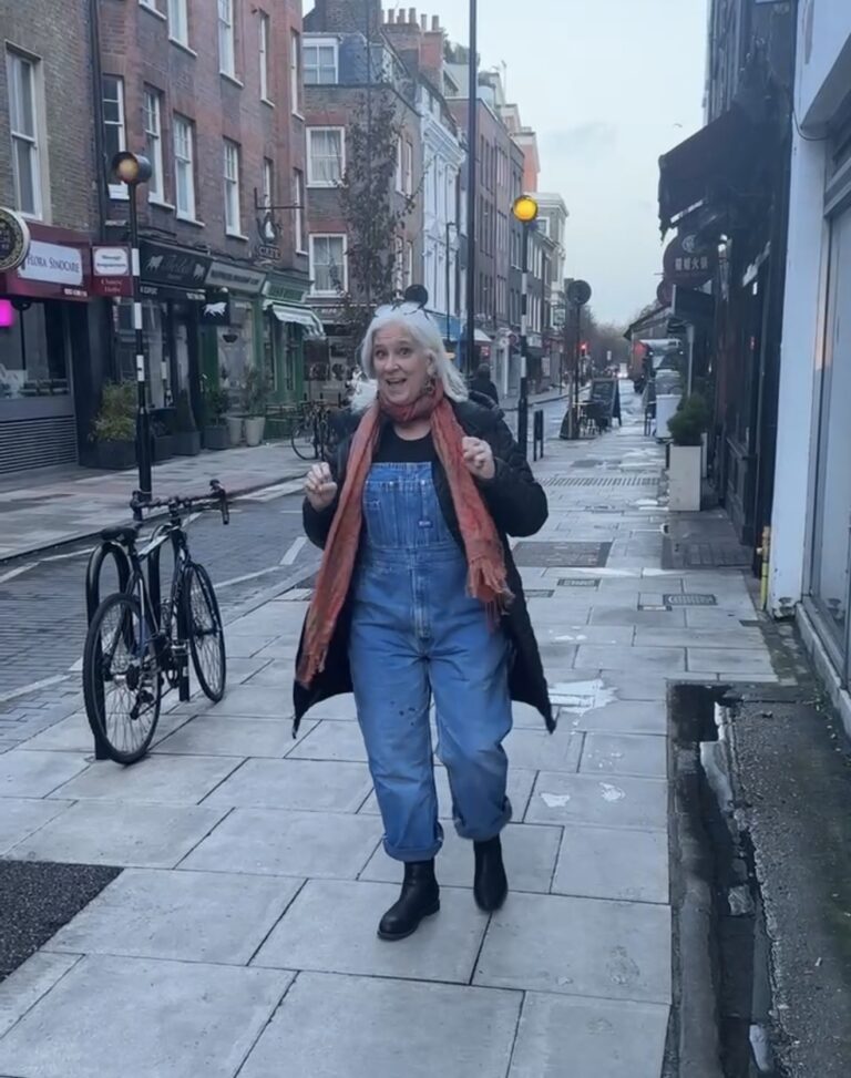 Jules Morris walking a London street in vintage denim overalls, a black coat and a coral scarf, shop fronts either side