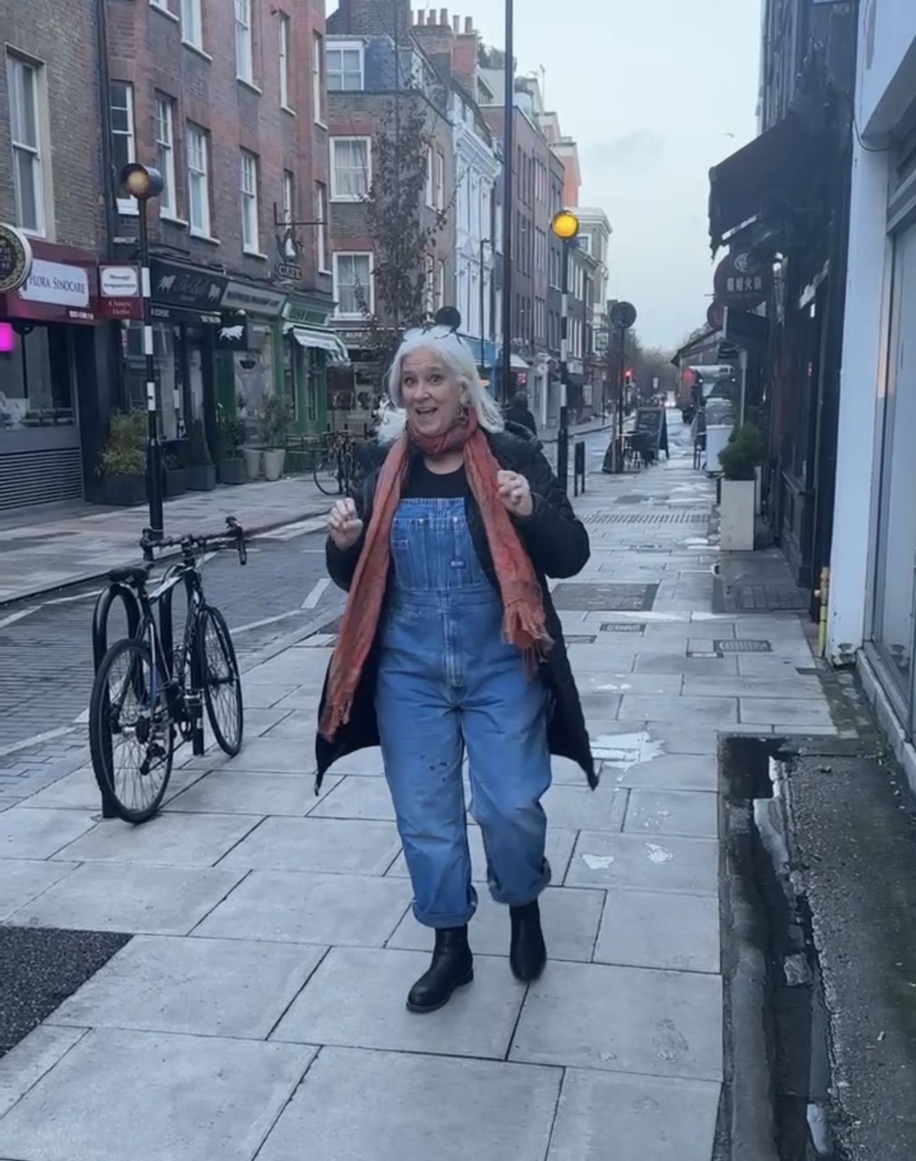 Jules Morris walking a London street in vintage denim overalls, a black coat and a coral scarf, shop fronts either side