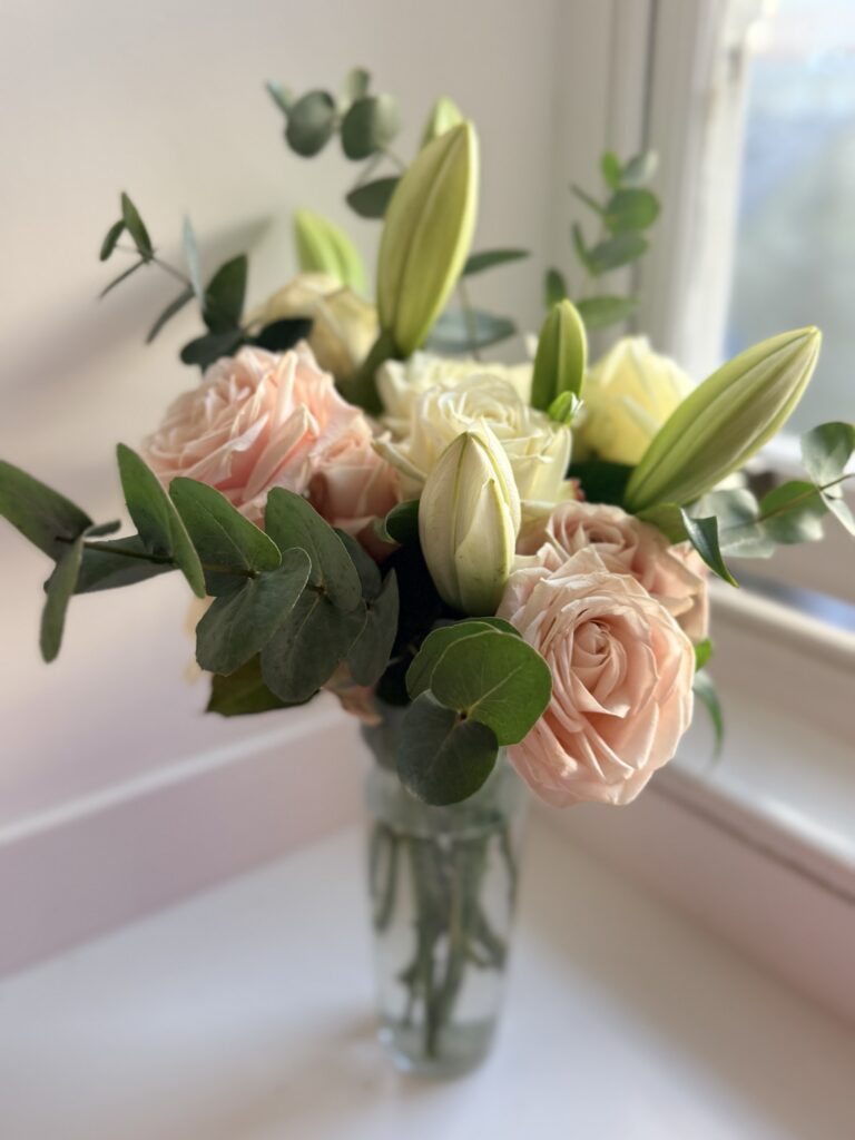 Arrangement of blush roses and lilies with eucalyptus in a glass vase on a windowsill