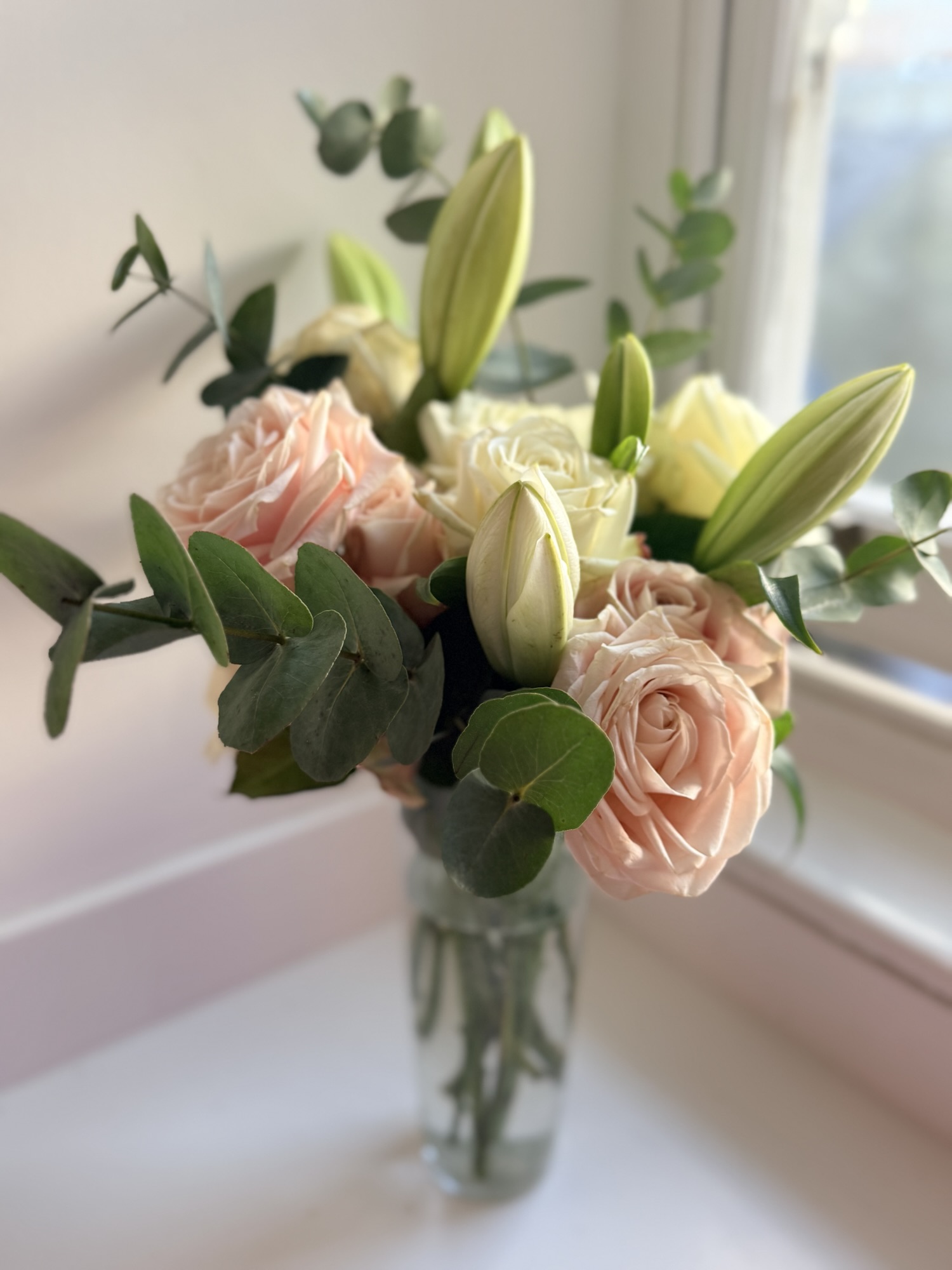 Arrangement of blush roses and lilies with eucalyptus in a glass vase on a windowsill