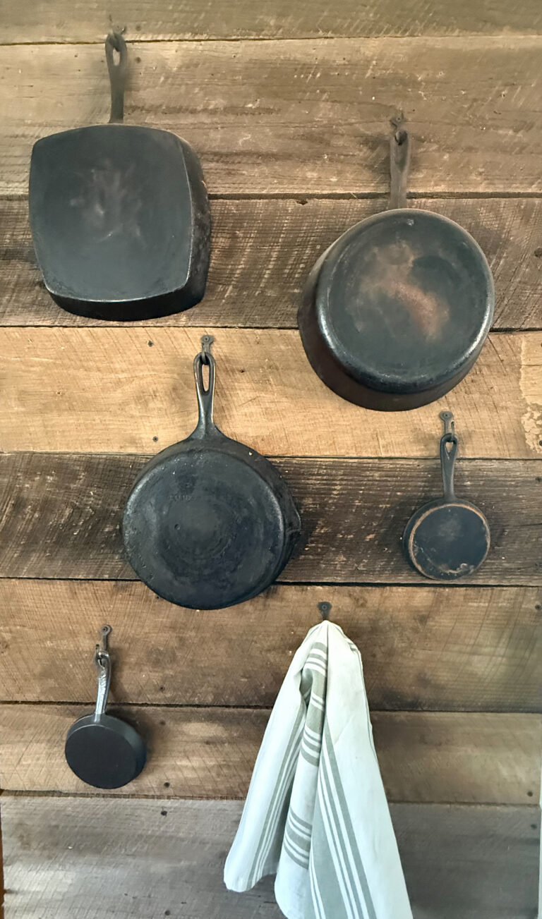 Five pieces of restored cast iron hanging from forged hooks on a reclaimed wood wall beside a striped kitchen towel