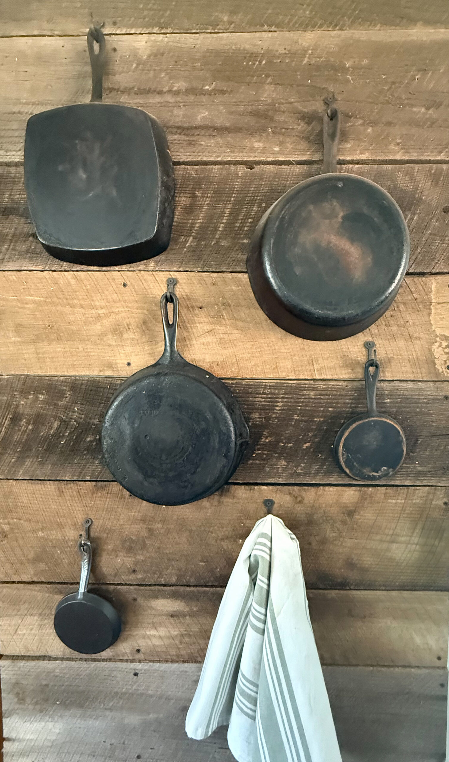 Five pieces of restored cast iron hanging from forged hooks on a reclaimed wood wall beside a striped kitchen towel
