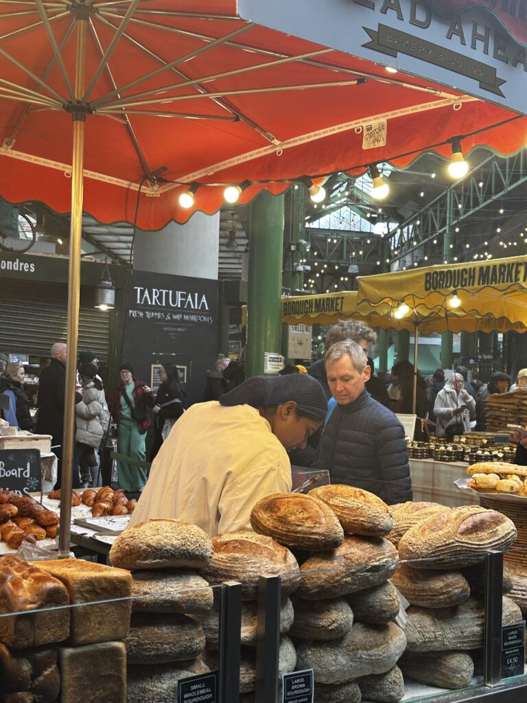 Stacks of round sourdough loaves and croissants at the Bread Ahead bakery stall under a red umbrella at Borough Market