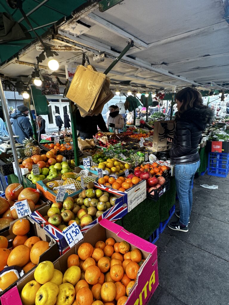 Outdoor London fruit stall with oranges, apples and pomegranates in Union Jack printed crates, a shopper choosing fruit