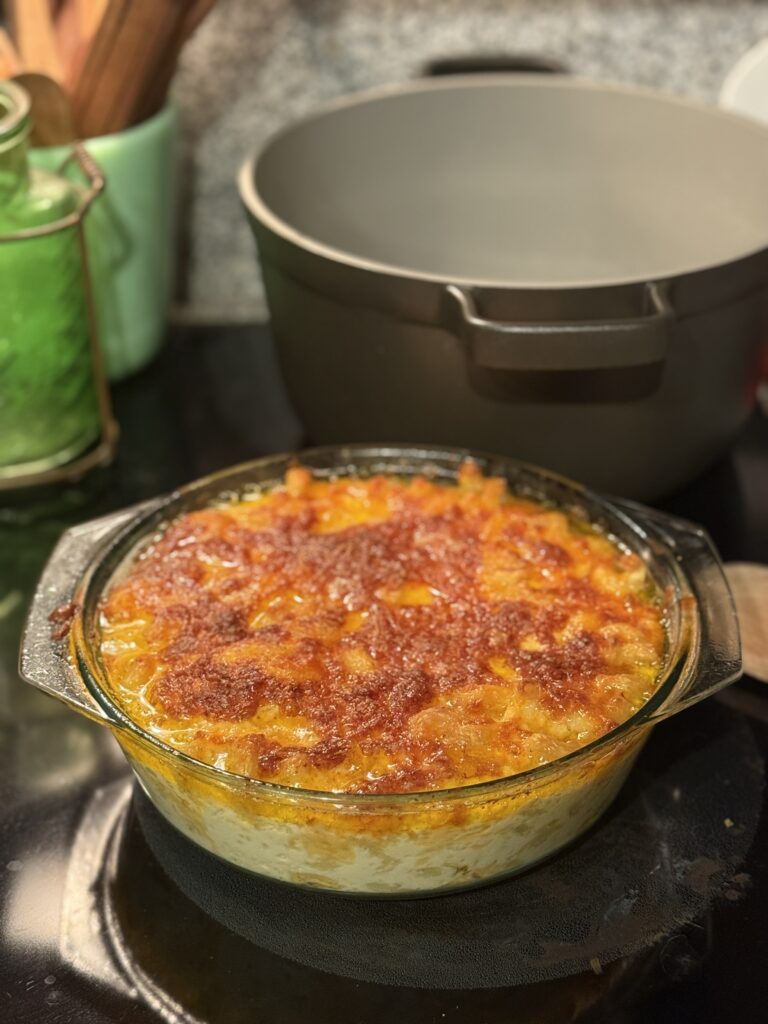 Round glass casserole of baked macaroni and cheese with a deeply browned bubbling top, cooling on the stovetop