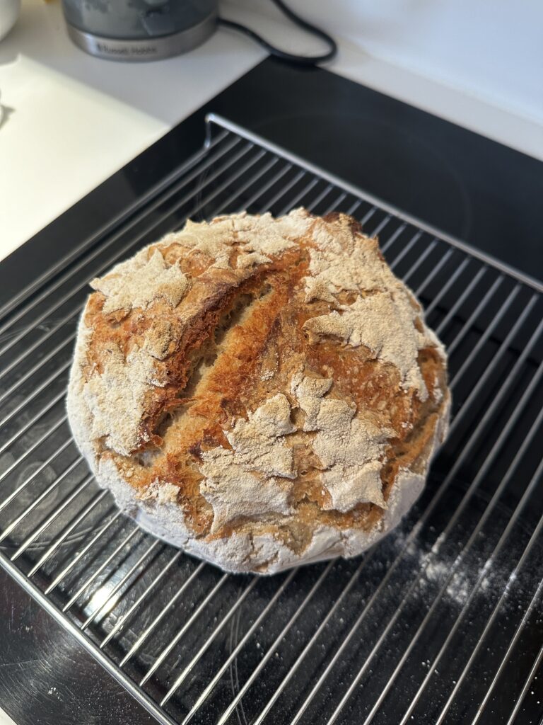 Round sourdough loaf with a heavily floured crackled crust and a deep open ear, cooling on a rack