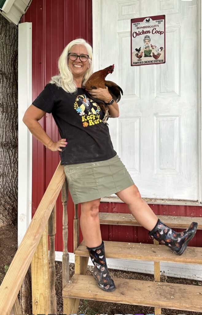 Jules Morris standing on the coop steps holding a red rooster, beside a sign reading BombDiggity Chicken Coop, Pampered Chickens Live Here