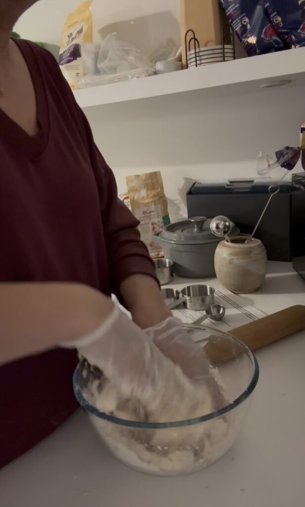 Jules Morris mixing sourdough by hand in a glass bowl on the counter of the London flat, flour and measuring cups beside her