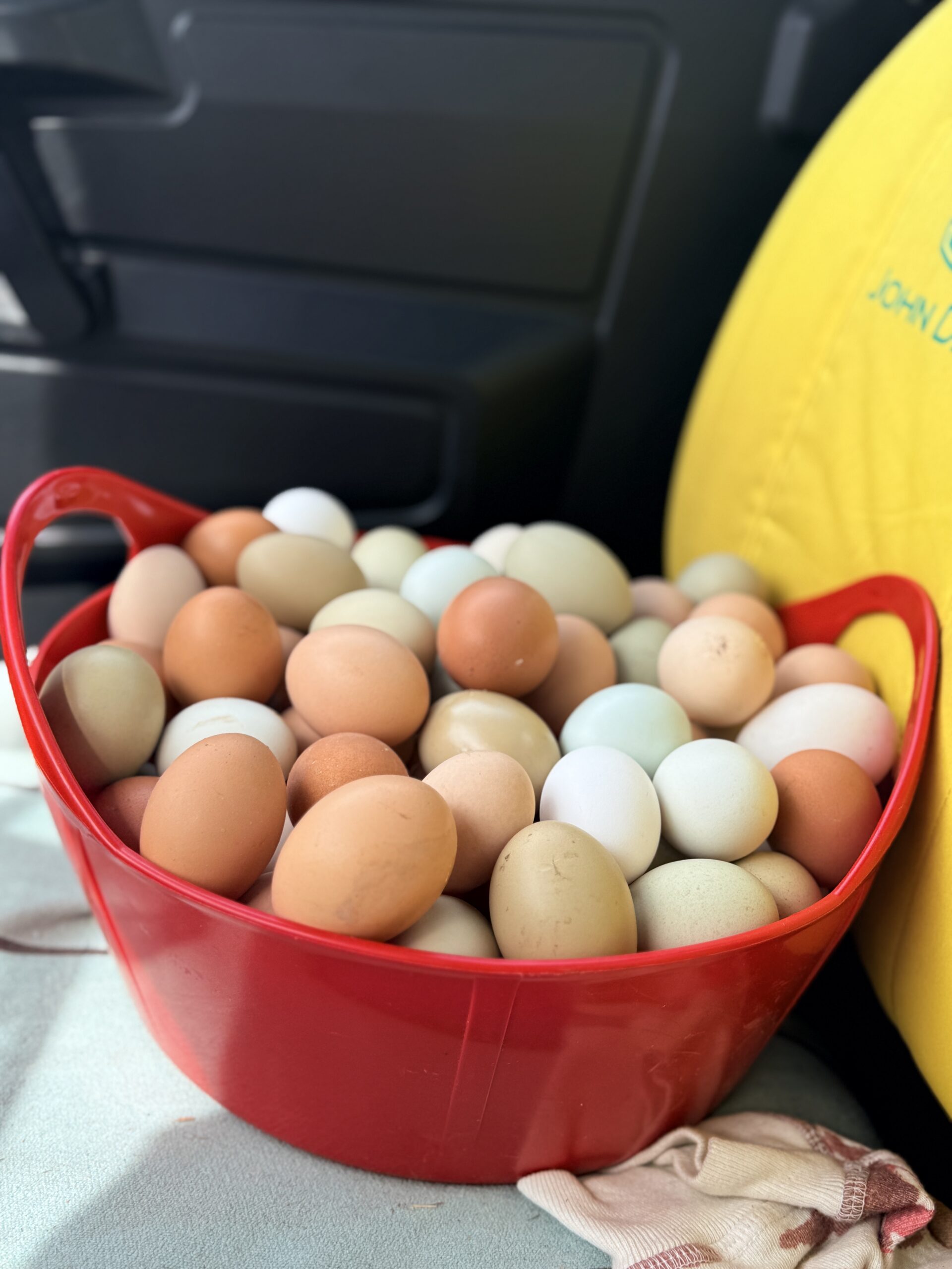 Red tub heaped with farm fresh chicken eggs in brown, olive green, pale blue, cream and white, sitting on a tractor seat