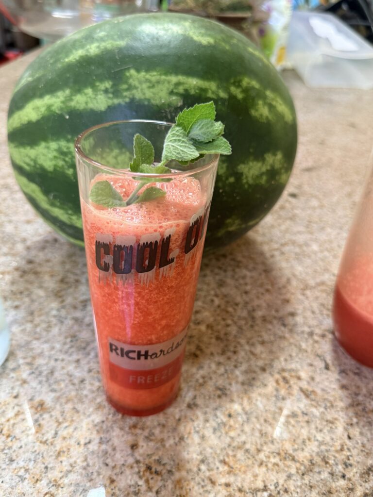 A watermelon mint cooler in a tall glass topped with fresh mint, beside a whole watermelon on the kitchen counter