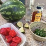 Ingredients for a watermelon mint cooler laid out on the counter: cubed watermelon, limes, raw honey, sparkling water and fresh mint in a stoneware bowl