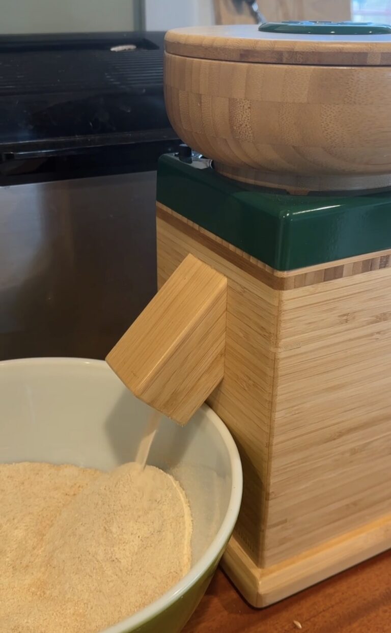 Bamboo bodied NutriMill grain mill on the counter with freshly milled flour pouring from the chute into a bowl