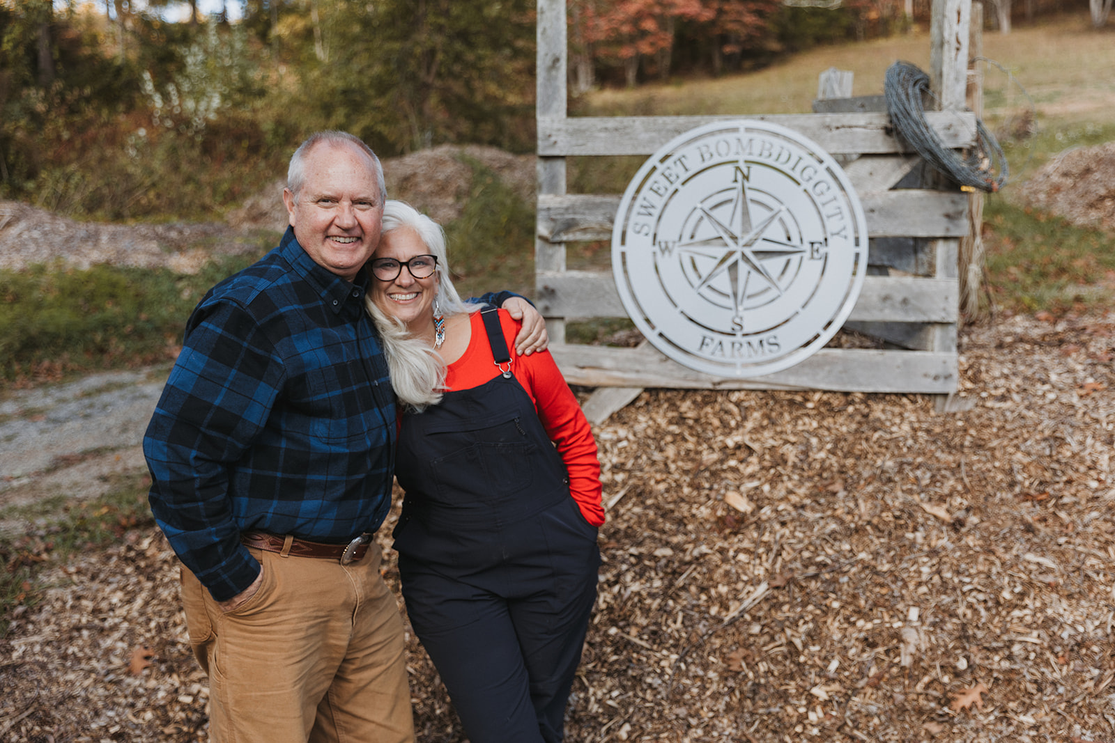 Lane and Jules Morris standing arm in arm in front of the farm gate and its compass sign in the fall