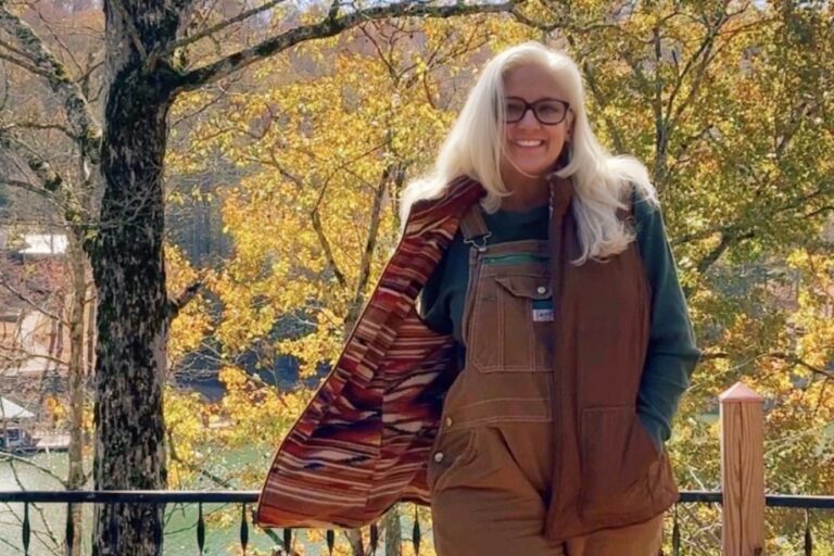 Jules Morris on the lake deck in brown bib overalls and a vest with a striped lining, fall leaves behind her