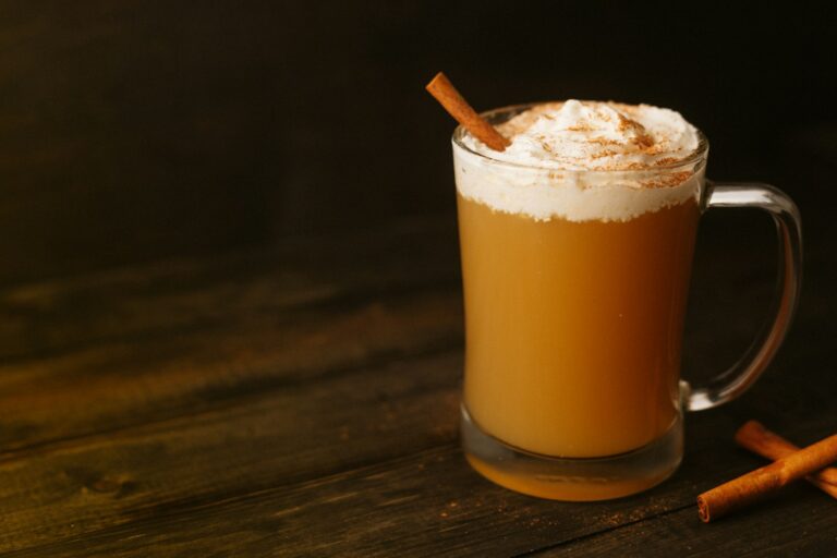 A glass mug of hot apple cider topped with foam and a cinnamon stick on a dark wooden table