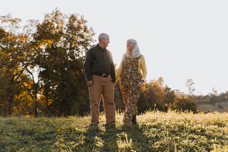 Lane and Jules Morris holding hands and smiling at each other in a sunlit field, with fall trees and rolling hills behind them
