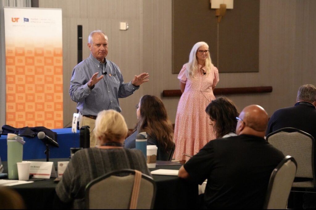 Lane and Jules Morris leading a workshop, Lane speaking to participants seated at round tables while Jules stands beside him