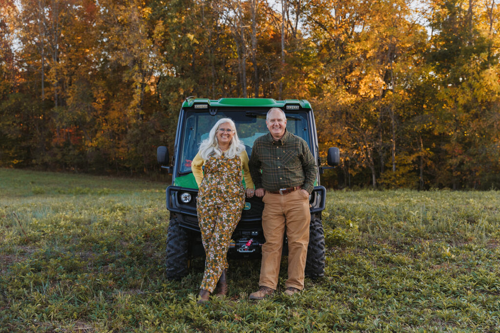 Lane and Jules Morris smiling as they lean on the front of their green John Deere XUV in the field, with fall trees behind them