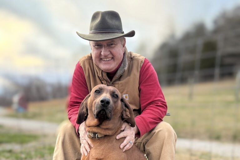 Lane Morris in a felt hat and Carhartt vest crouching with his arms around a redbone hound on the farm
