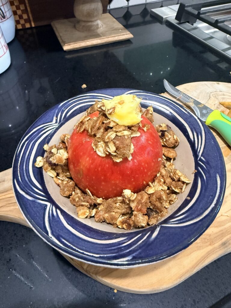 Red apple packed with oat, brown sugar and pecan filling and topped with butter in a blue and white ceramic apple baker, ready for the oven