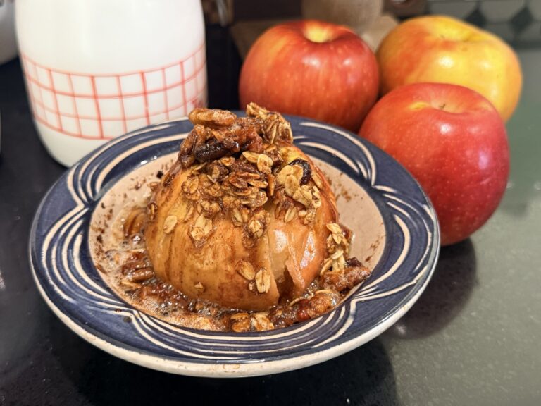 Stuffed baked apple with a golden oat and pecan filling in a blue and white ceramic apple baker, with fresh red apples behind it