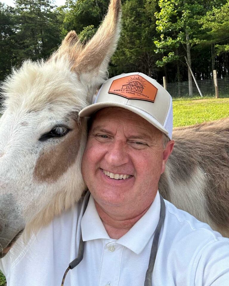 Lane Morris grinning in a ball cap with a fluffy donkey pressed cheek to cheek against him